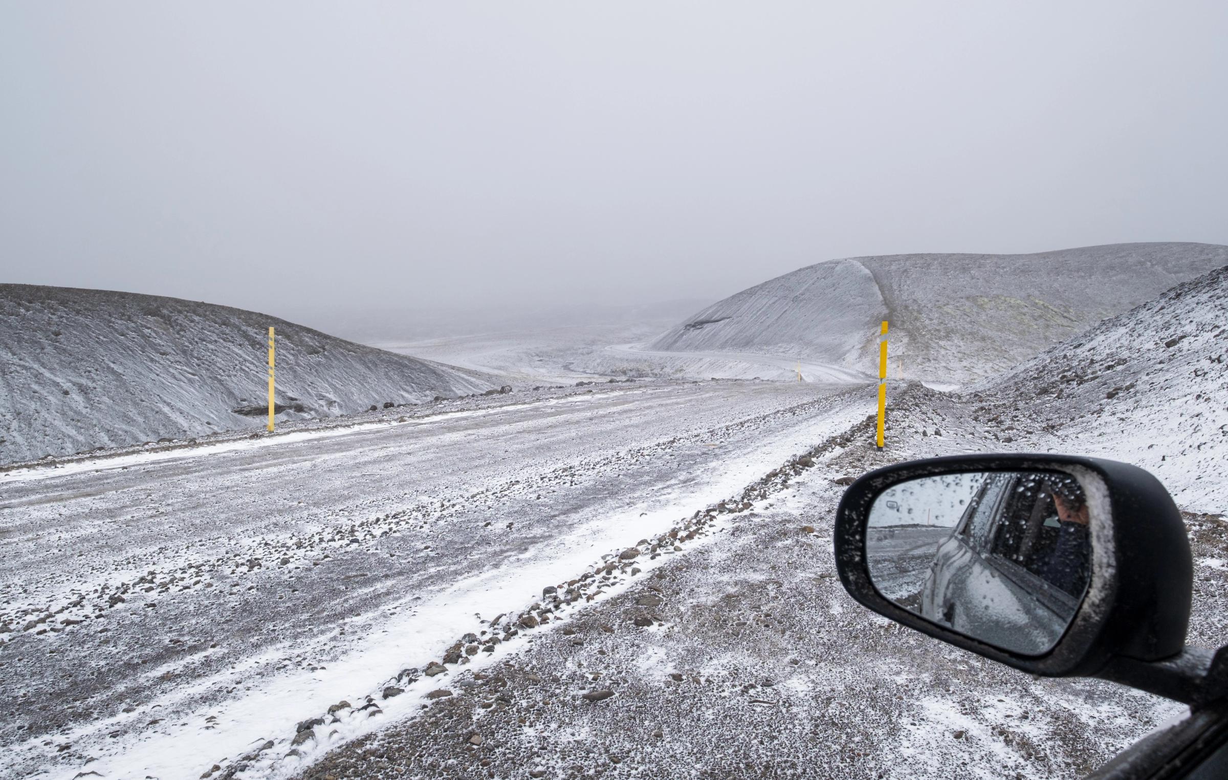 Snowy gravel road winding through desolate hills under a grey sky, seen from a car's side mirror, with yellow marker poles.