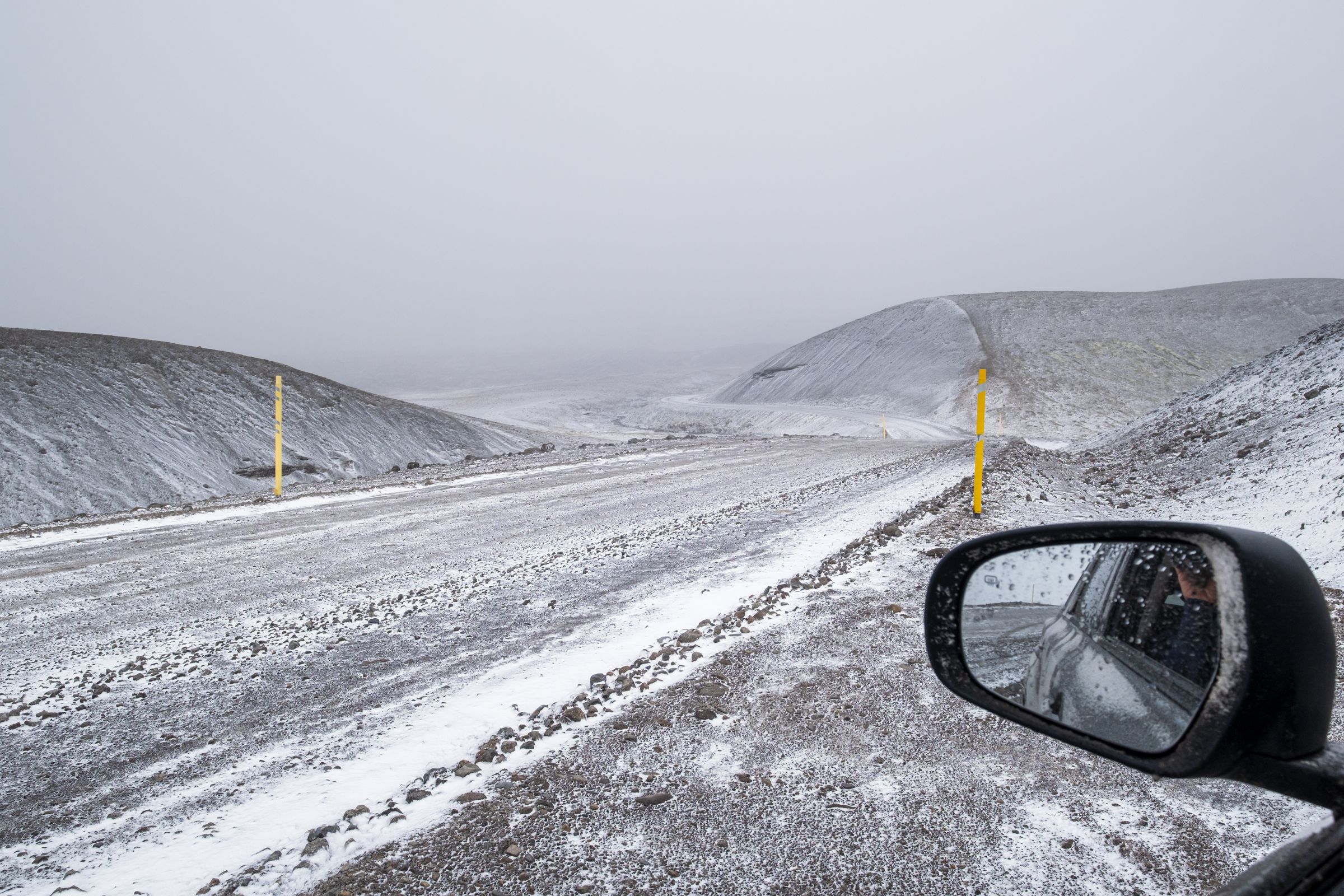 Snowy gravel road winding through desolate hills under a grey sky, seen from a car's side mirror, with yellow marker poles.