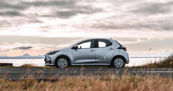 A silver hatchback car parked by a scenic roadside with water, mountains, and a cloudy sky in the background.