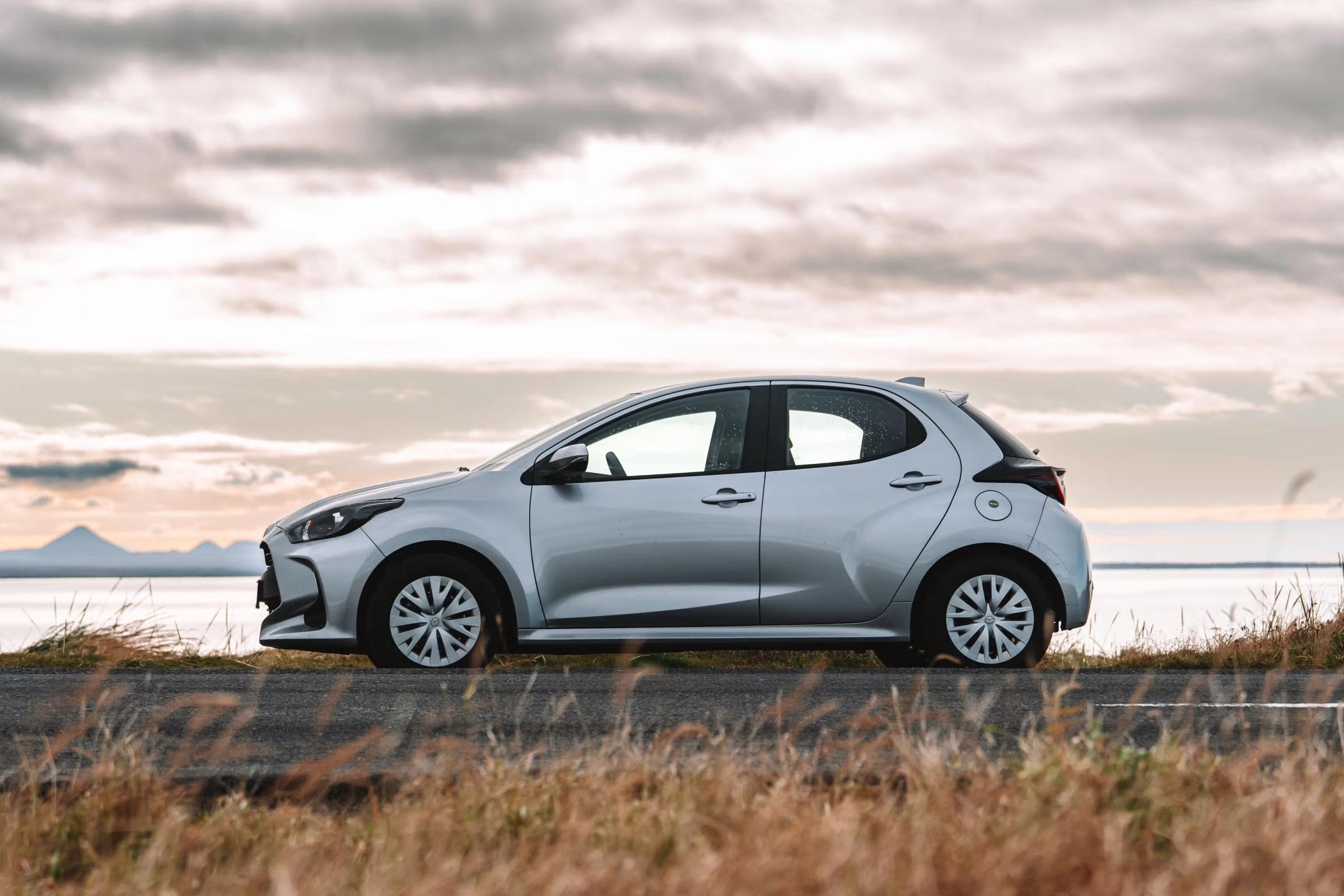 Toyota Yaris plateado, modelo hatchback, estacionado al costado de la carretera con vista al agua bajo un cielo nublado.