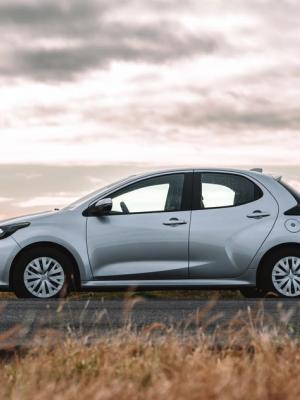A silver hatchback car parked by a scenic roadside with water, mountains, and a cloudy sky in the background.