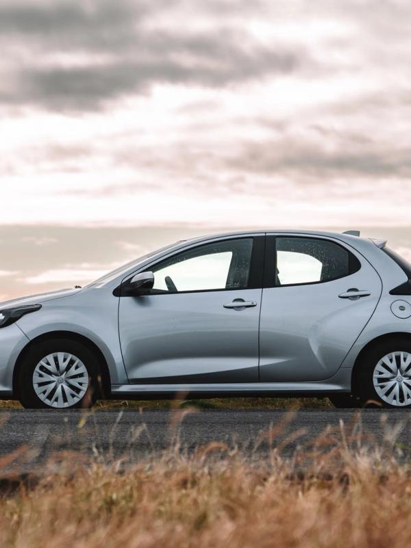 A silver hatchback car parked by a scenic roadside with water, mountains, and a cloudy sky in the background.