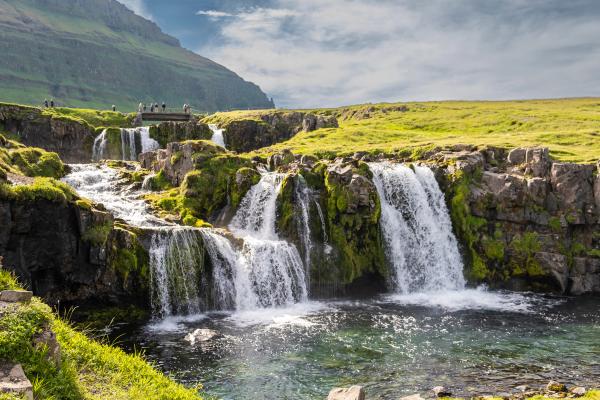 Cascading waterfall over mossy rocks with a bridge and people above.
