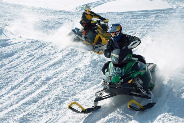 Two snowmobilers ride through deep snow, kicking up powder.