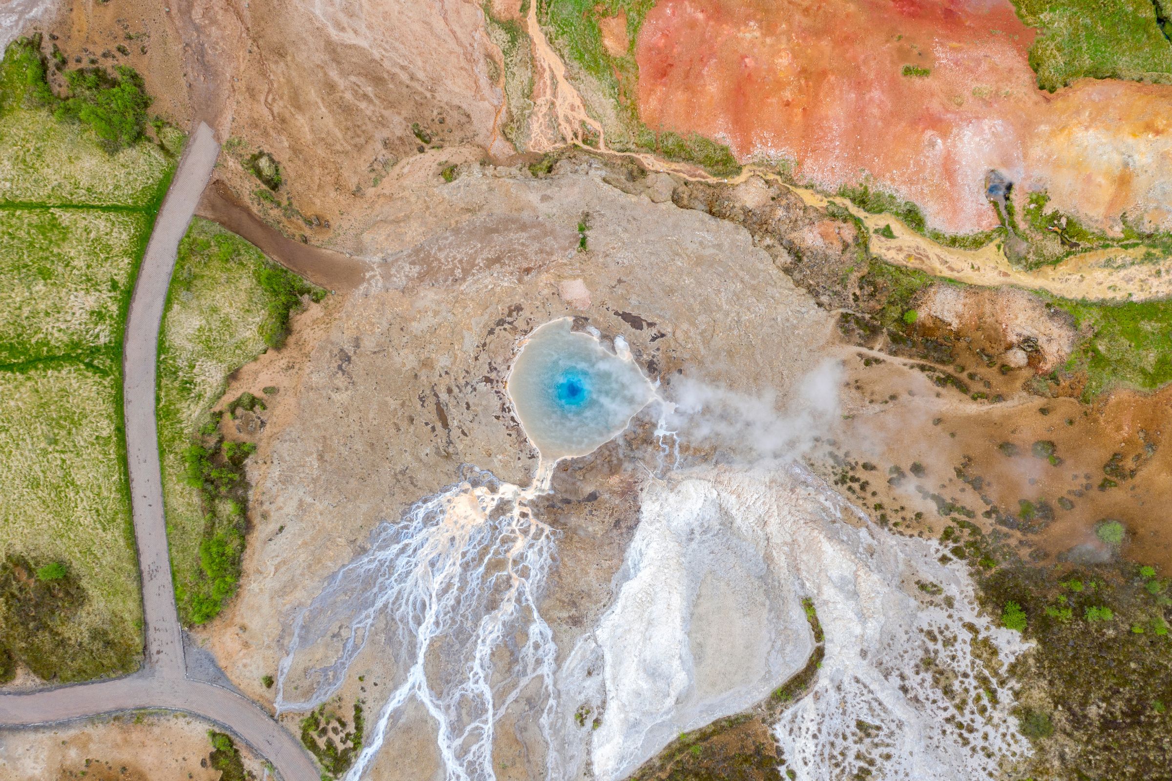 Aerial view of a vibrant blue hot spring, steaming and surrounded by white mineral deposits, colorful geothermal ground, and a winding path.