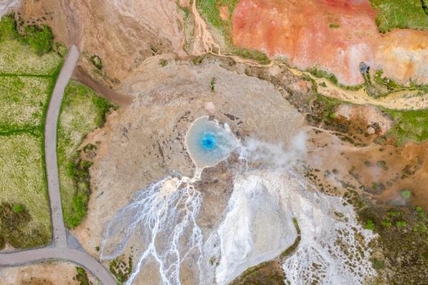 Aerial view of a bright blue geyser pool steaming amidst white mineral deposits and colorful geothermal terrain.
