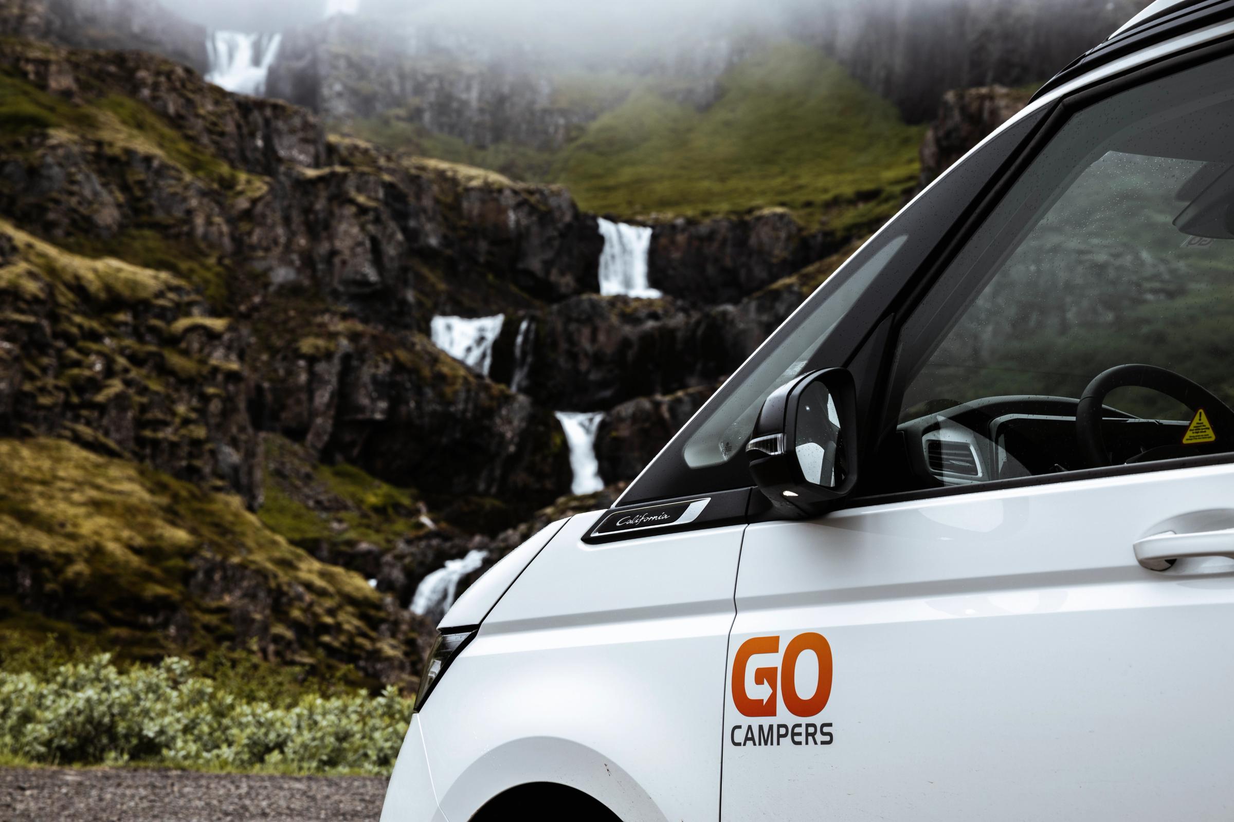 Side view of a white Go Campers van parked before a green, rocky mountain with multiple cascading waterfalls.