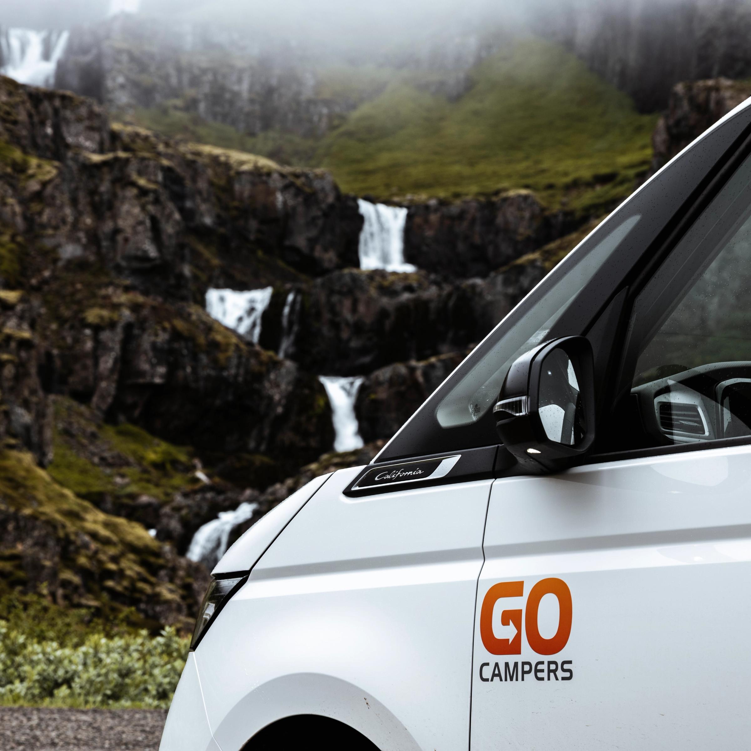 Side view of a white Go Campers van parked before a green, rocky mountain with multiple cascading waterfalls.