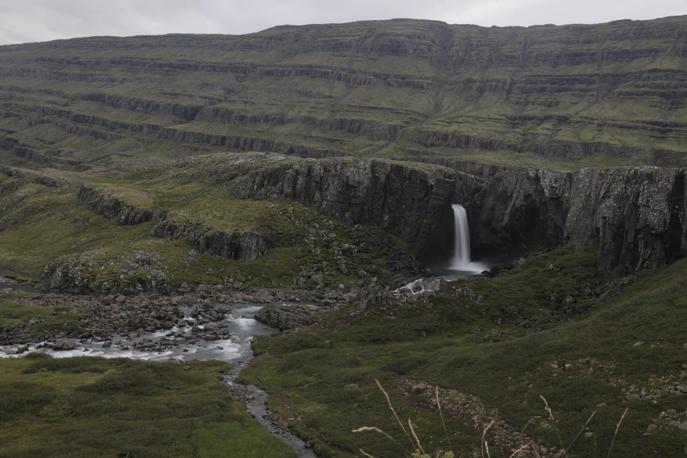 A waterfall cascades into a rocky canyon, with a stream flowing through a green, rugged valley and layered hills under an overcast sky.