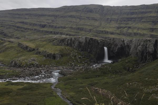 A waterfall cascades into a rocky canyon, with a stream flowing through a green, rugged valley and layered hills under an overcast sky.