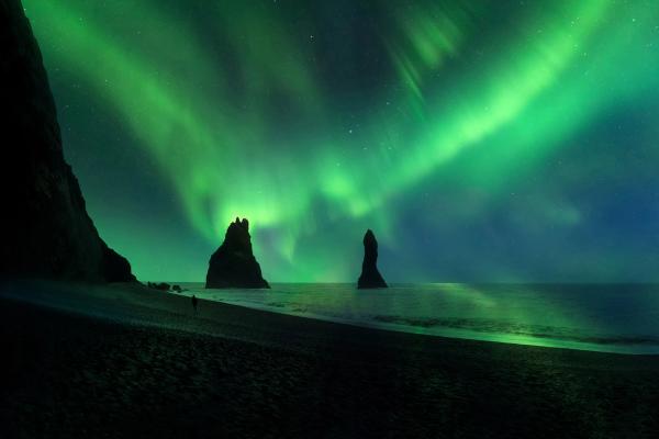 Green Northern Lights illuminate a black sand beach with sea stacks and a lone figure.