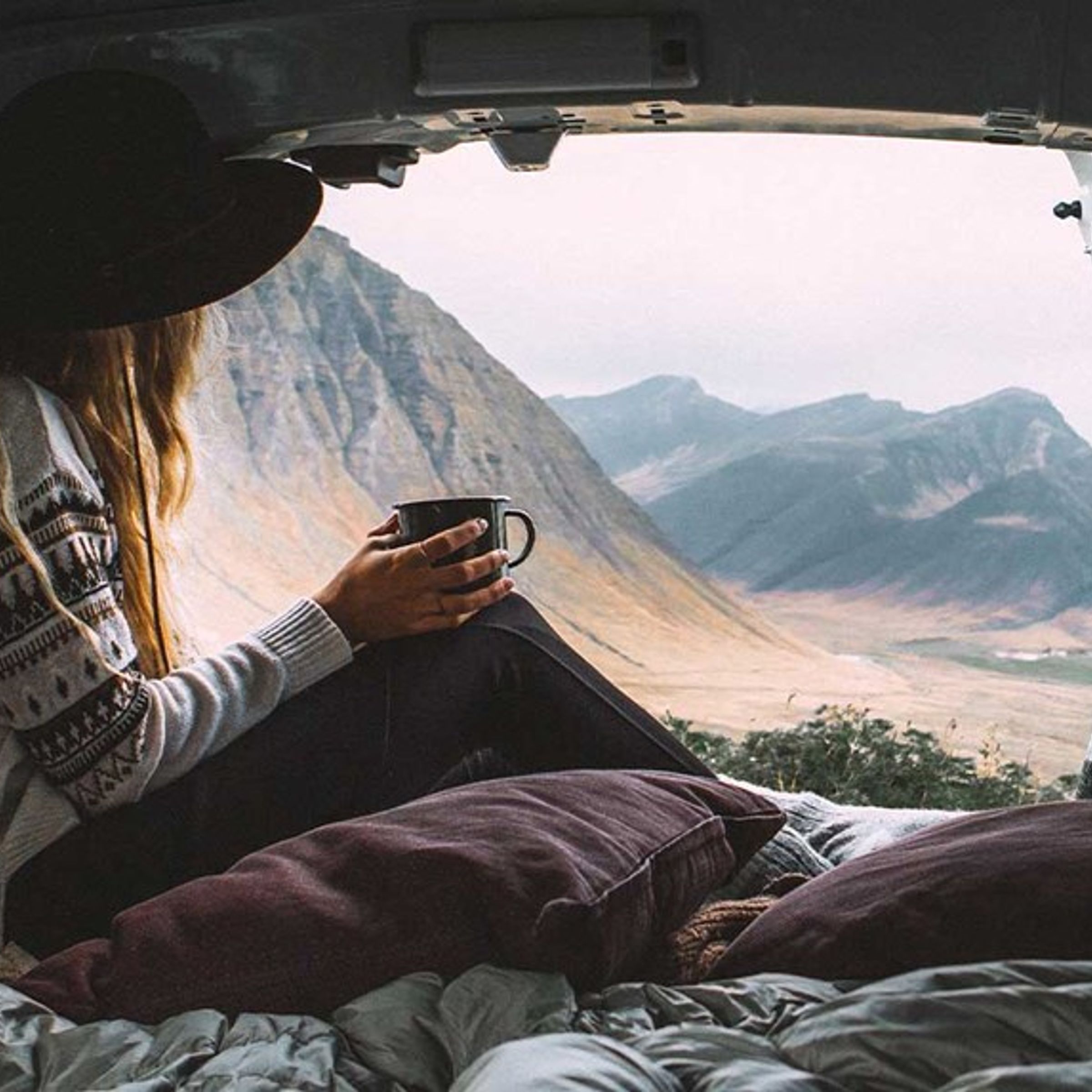 A woman in a hat sits in the back of a van, holding a mug and looking out at a mountainous landscape.