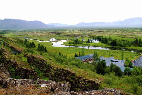 Green valley landscape with a winding river, distant mountains, buildings, and moss-covered rock formations in the foreground.