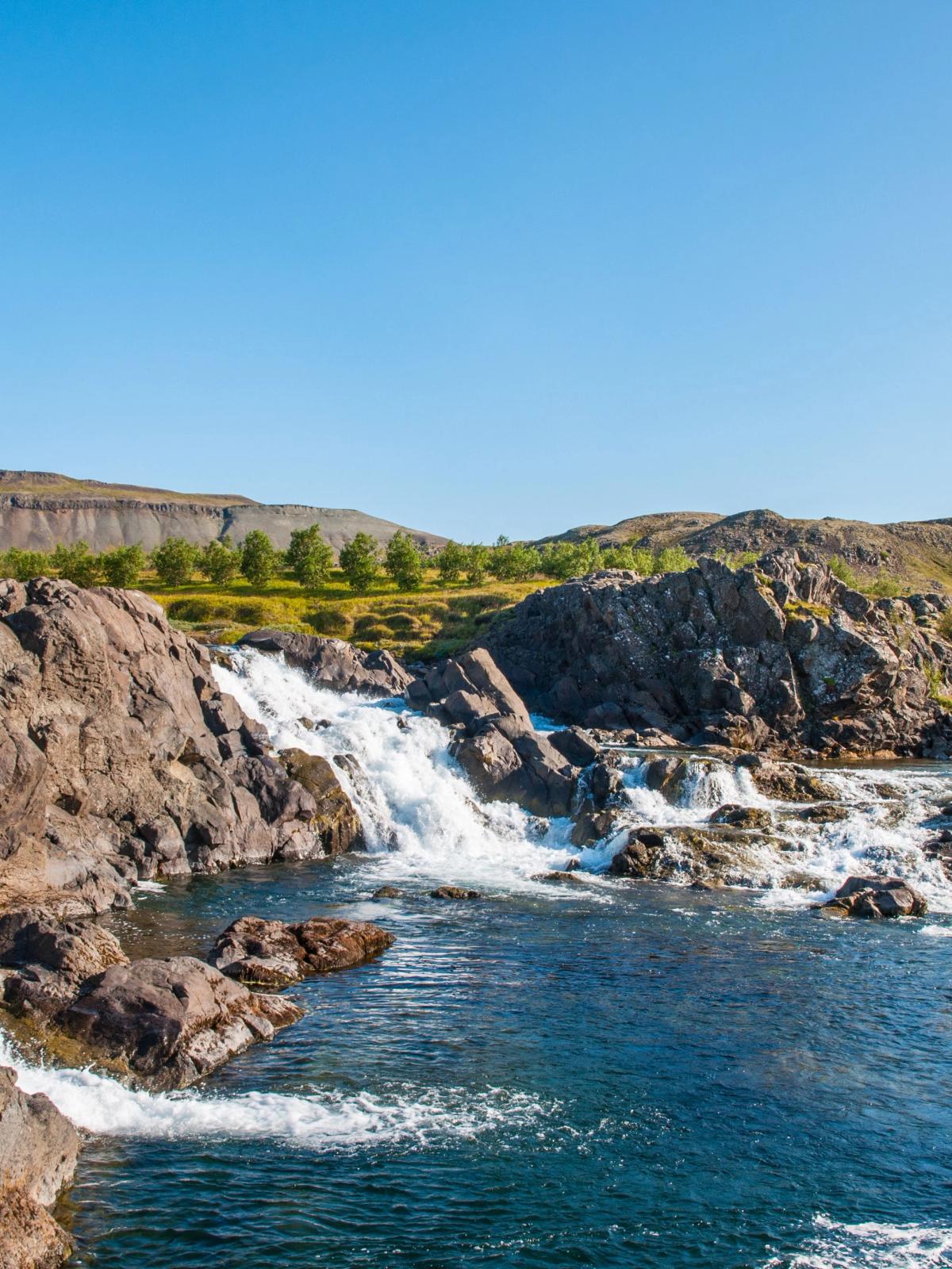 A waterfall cascades over dark rocks into a blue river, with green hills under a clear blue sky.