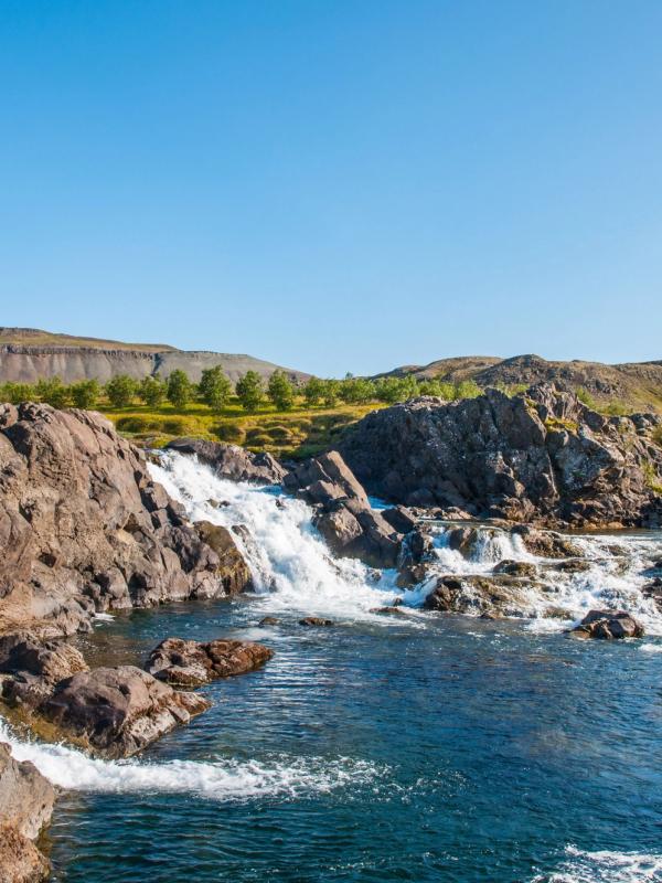 A waterfall cascades over dark rocks into a blue river, with green hills under a clear blue sky.