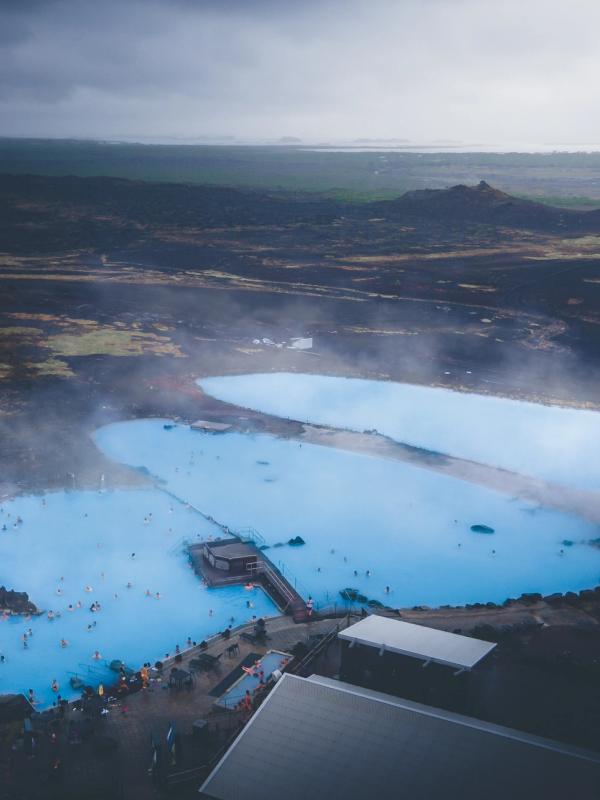 Aerial view of the Blue Lagoon in Iceland, showing milky blue thermal pools filled with people, surrounded by dark volcanic terrain, with steam rising.