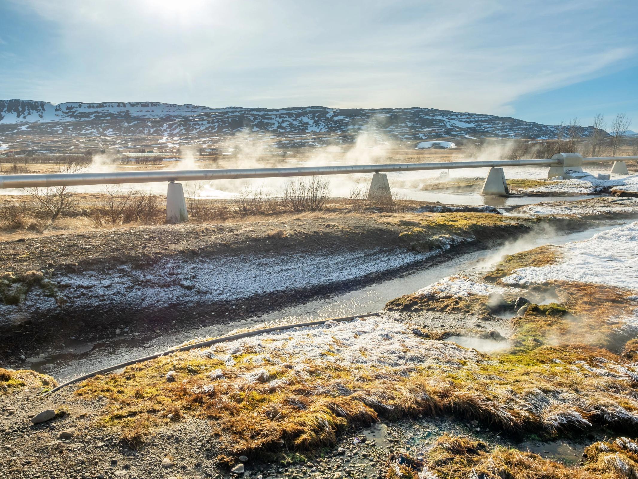 Una gran tubería plateada cruza un arroyo humeante en un paisaje nevado bajo un cielo soleado.