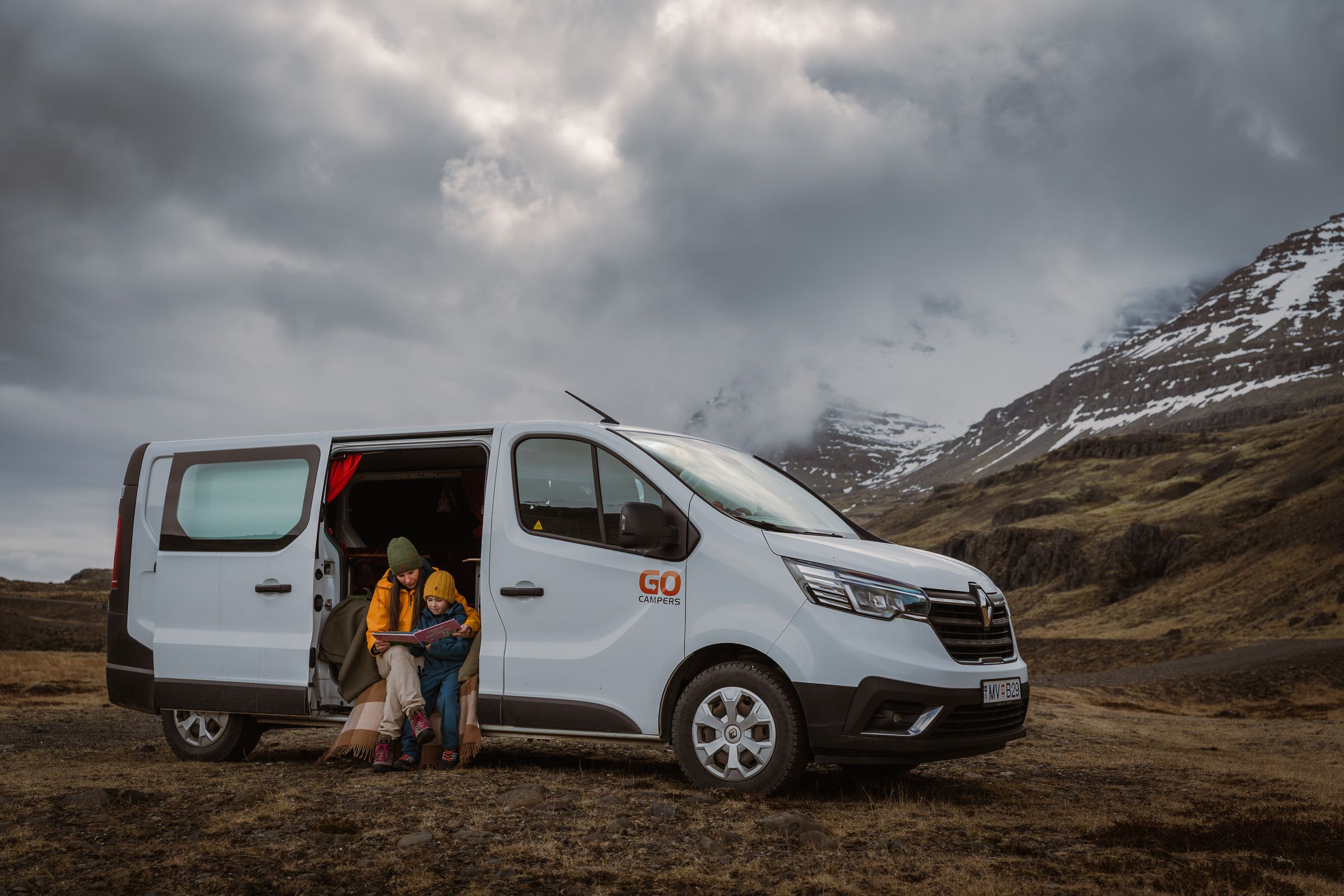 An adult and child sit in the open side door of a white "GO Campers" van, looking at a map against a cloudy mountain backdrop.