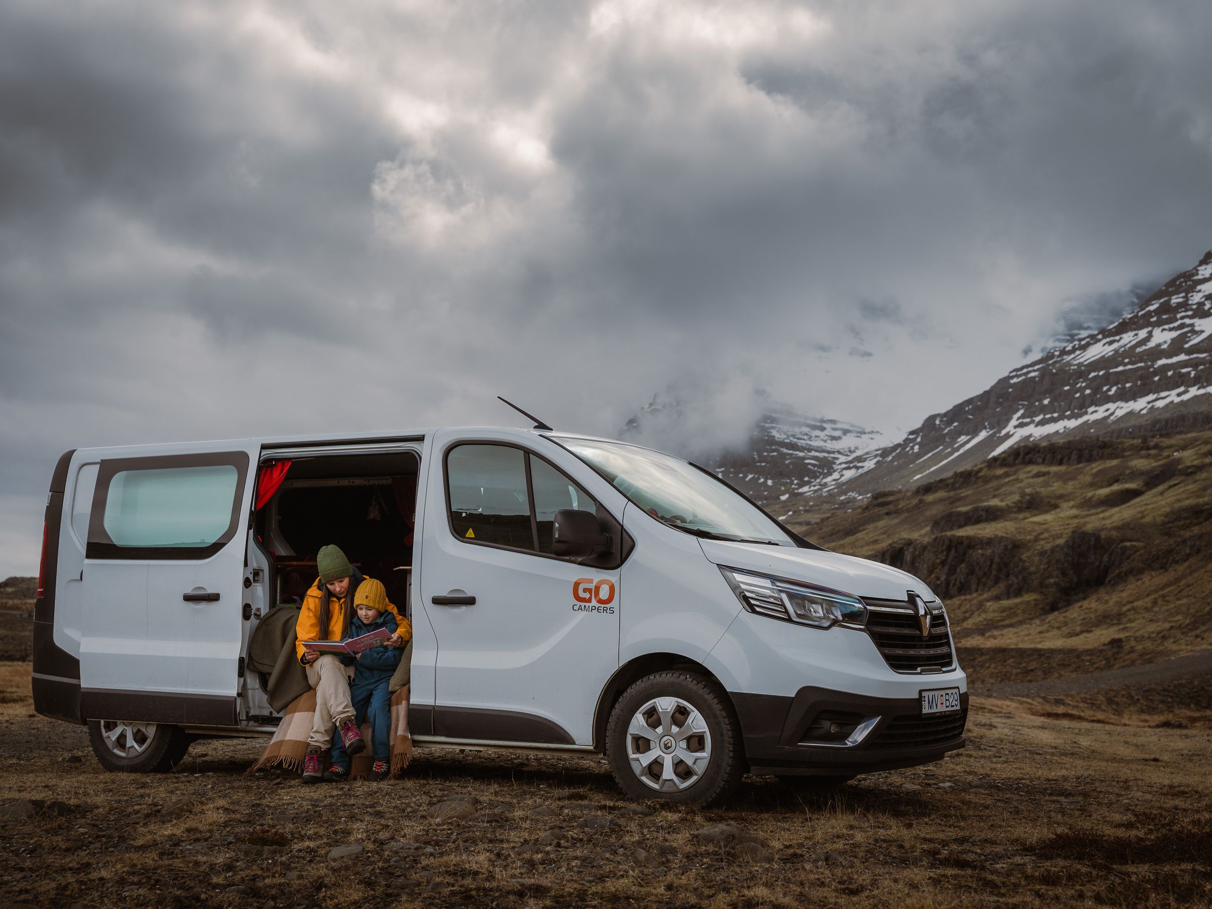 An adult and child sit in the open side door of a white "GO Campers" van, looking at a map against a cloudy mountain backdrop.