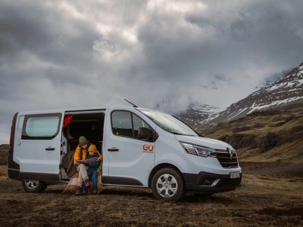 An adult and child sit in the open side door of a white "GO Campers" van, looking at a map against a cloudy mountain backdrop.