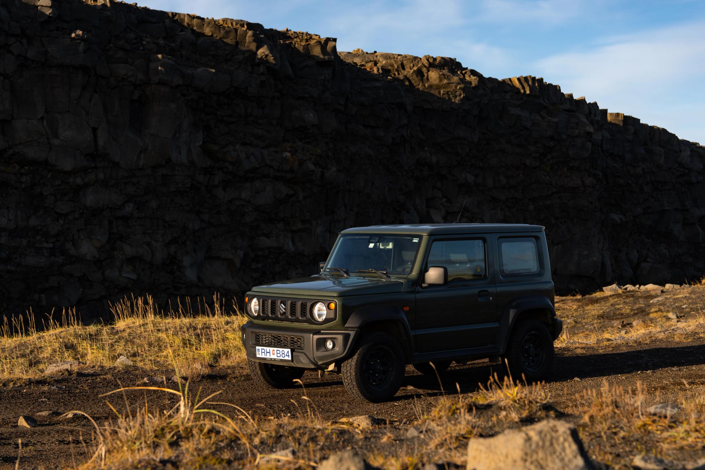 Suzuki Jimny 4x4 Mietwagen mit Schaltgetriebe in Island, geparkt auf einem unbefestigten Weg unterhalb einer dunklen Felswand