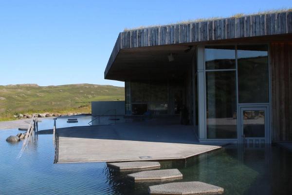 A modern building with a green roof and large windows sits next to a pool with stepping stones, set in a green, hilly landscape under a clear blue sky.