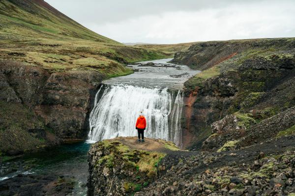 A person in an orange jacket stands on a cliff edge, looking at a large waterfall in a rugged, green landscape.