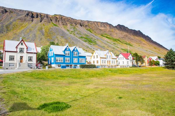 A row of colorful houses sits at the base of a large, grassy mountain with a green field in the foreground.