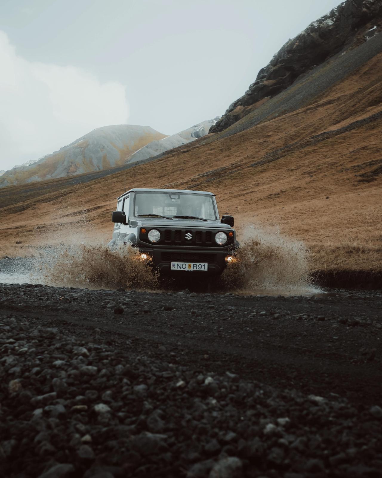 Un Suzuki Jimny gris oscuro salpica al pasar por un charco de barro en un camino pedregoso, con montañas al fondo.