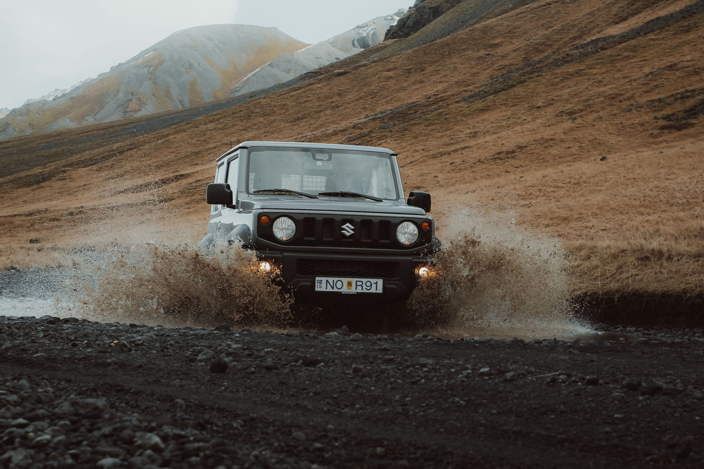 Un Suzuki Jimny gris oscuro salpica al pasar por un charco de barro en un camino pedregoso, con montañas al fondo.