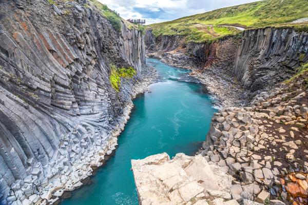 Un río de un turquesa intenso fluye por un profundo cañón, rodeado de altas y oscuras paredes de basalto columnar, coronadas por colinas verdes.
