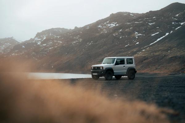 A silver Suzuki Jimny SUV parked on rocky terrain beside a lake, with dark, snow-dusted mountains in the background under an overcast sky.