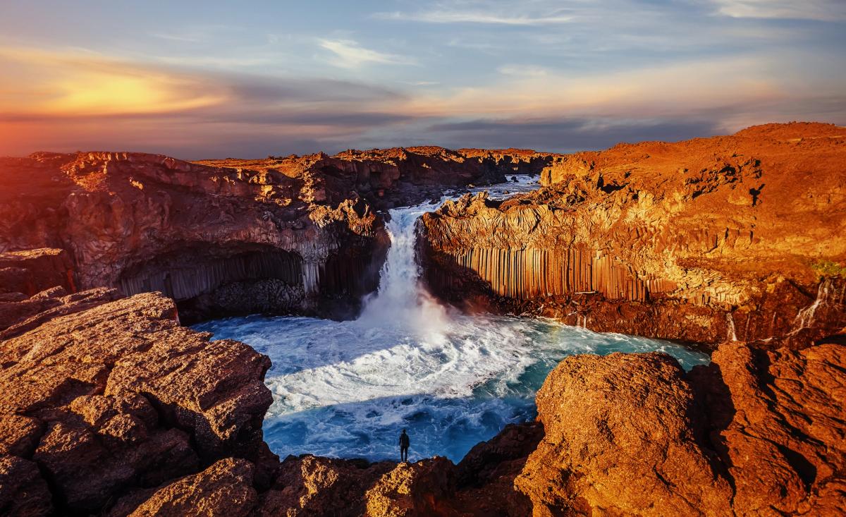 A person stands on a rocky cliff overlooking a powerful waterfall cascading between dramatic basalt columns under a colorful sky.