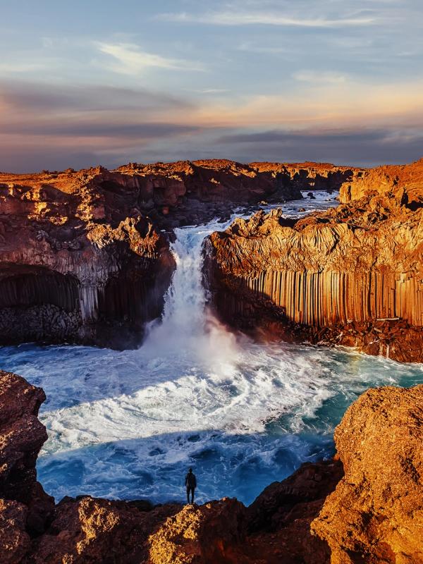 A person stands on a rocky cliff overlooking a powerful waterfall cascading between dramatic basalt columns under a colorful sky.