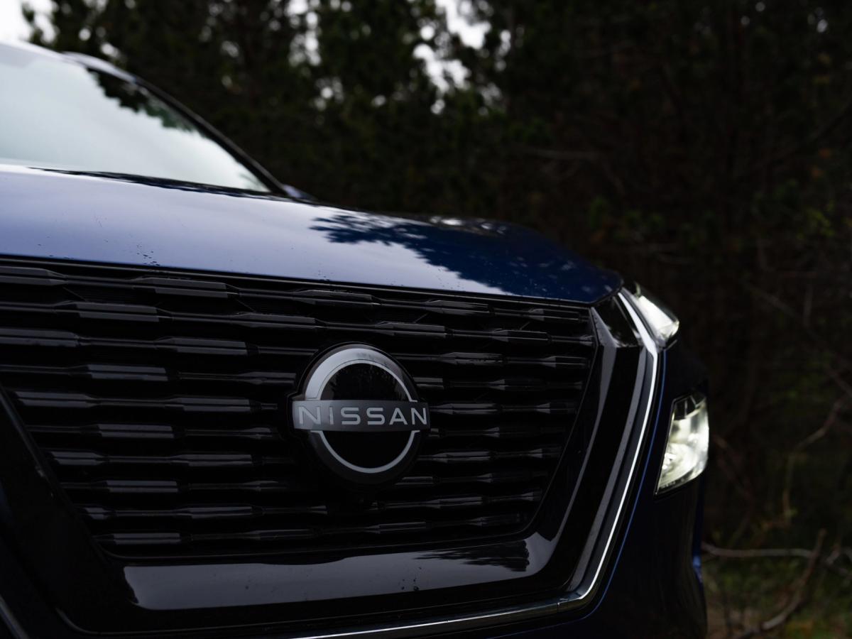 Detalle de la parrilla del X-Trail Primer plano de la parrilla delantera y el emblema de un Nissan X-Trail, con árboles al fondo