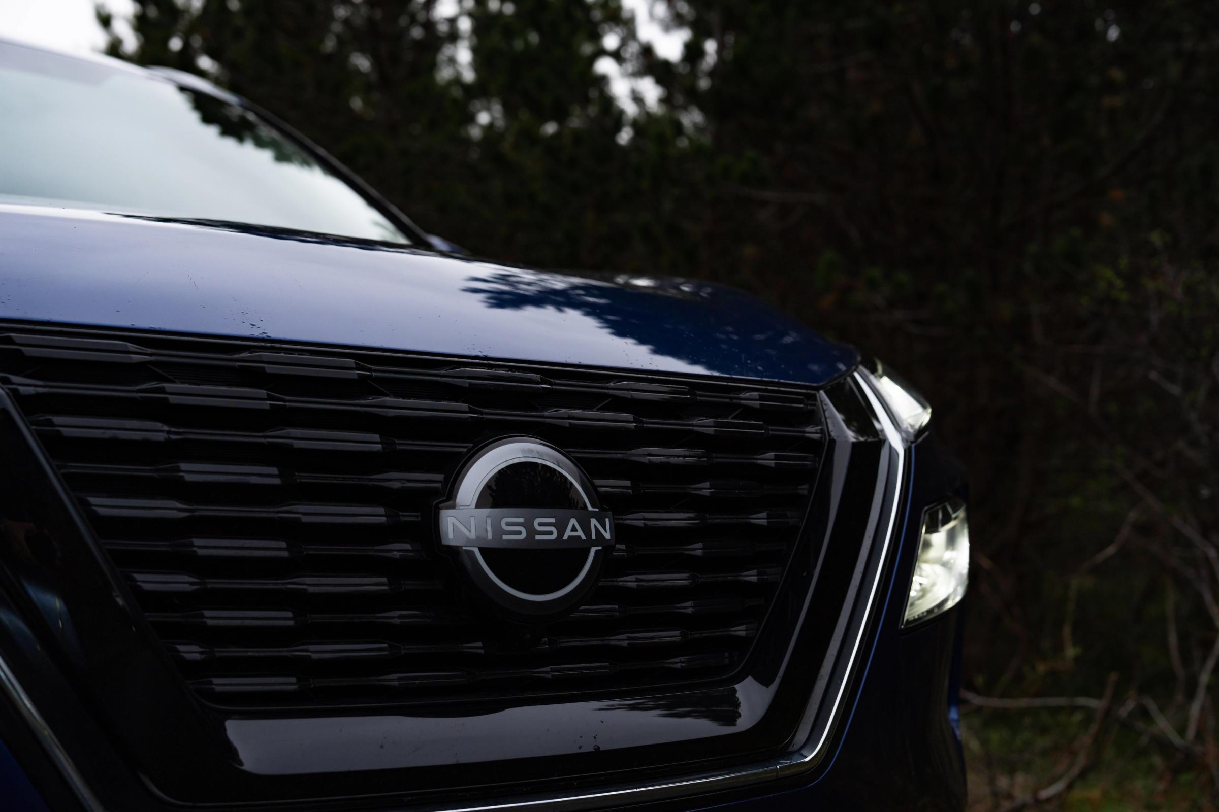 Detalle de la parrilla del X-Trail Primer plano de la parrilla delantera y el emblema de un Nissan X-Trail, con árboles al fondo