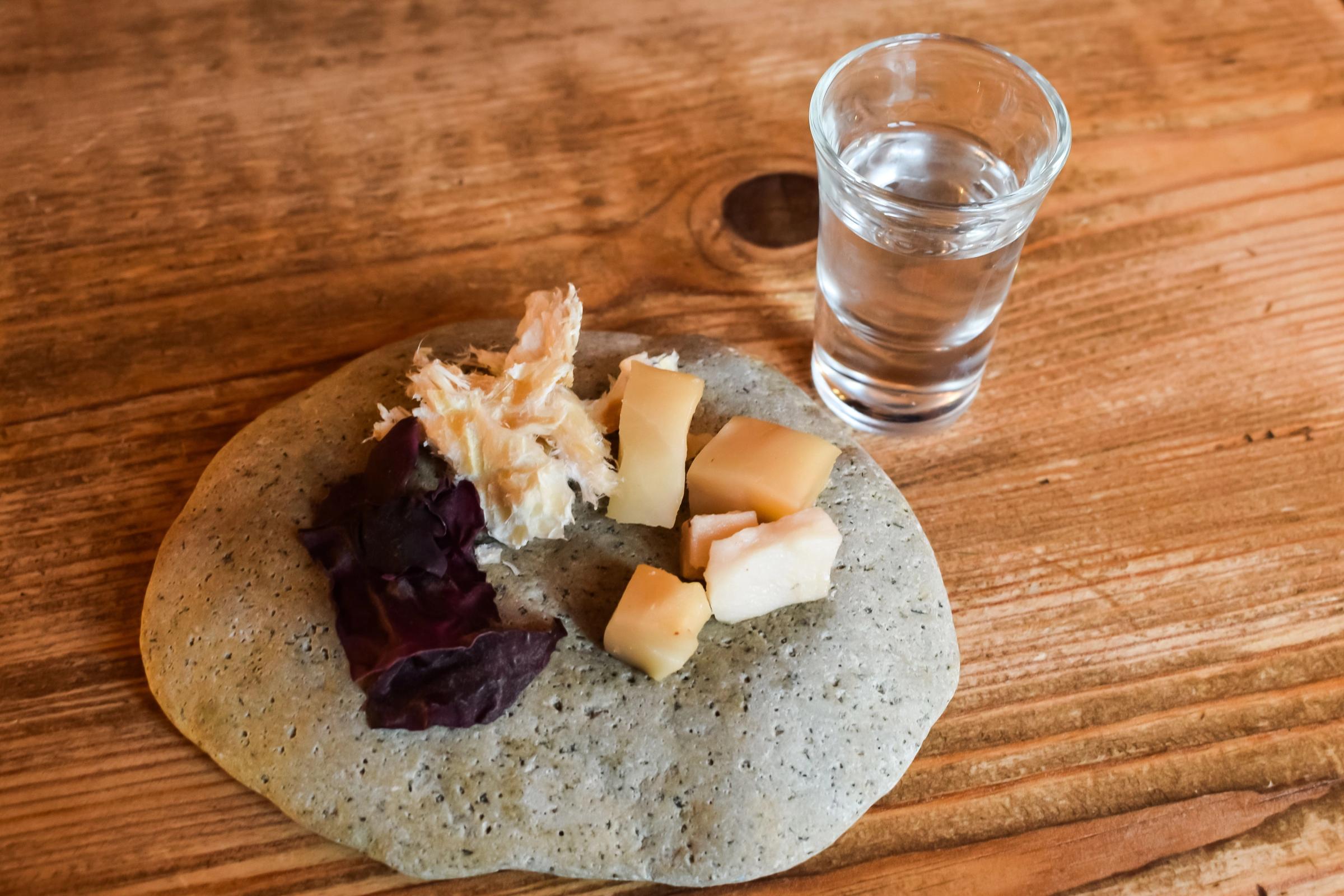 A grey platter with fermented shark, next to a shot glass of clear liquid on a wooden table.