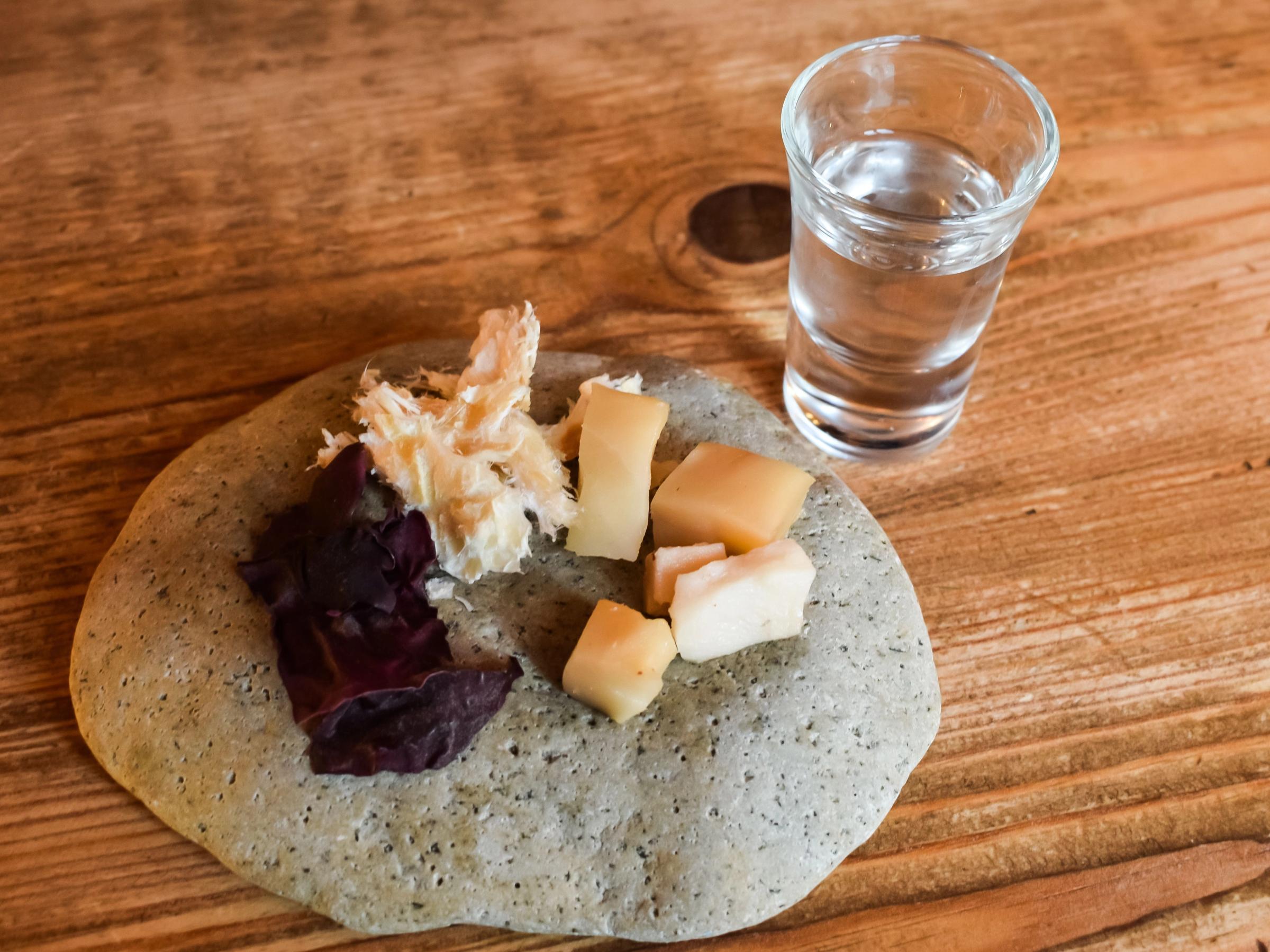 A grey platter with fermented shark, next to a shot glass of clear liquid on a wooden table.