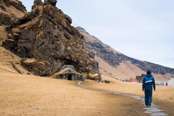 A person walks on a stone path through dry fields toward a turf house built into a rocky cliff.