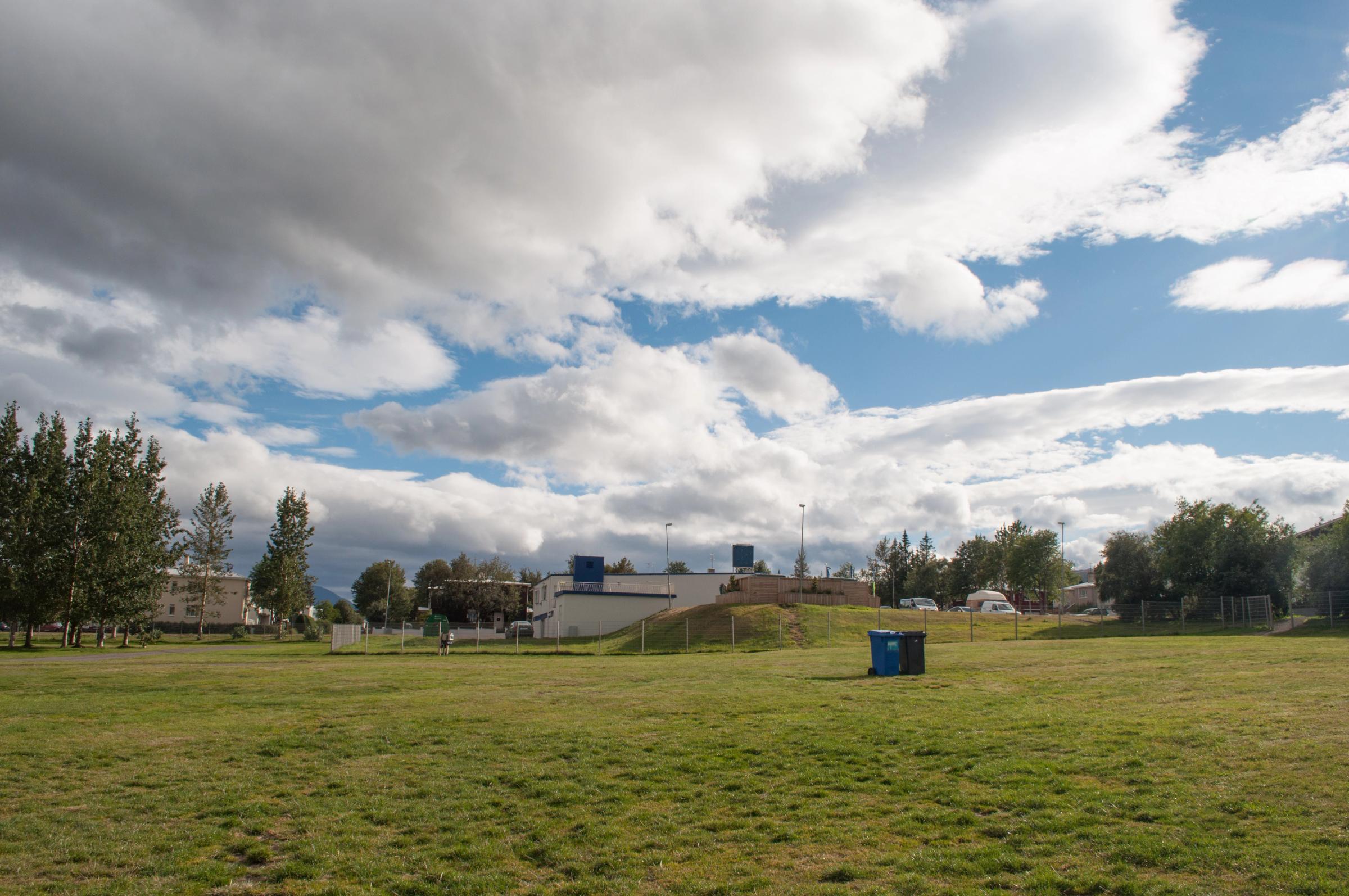 un amplio campo de hierba con un bote de basura azul en el medio bajo un cielo nublado.