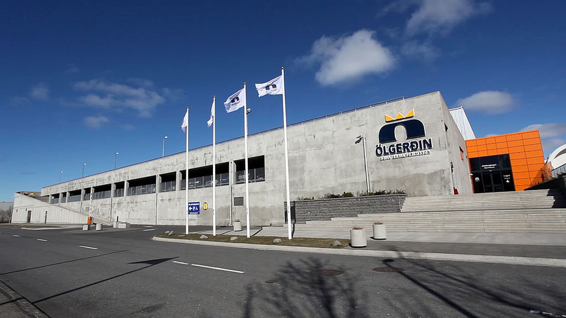Modern concrete building for Ölgerðin brewery with a crown logo, an orange section, and flags under a blue sky.