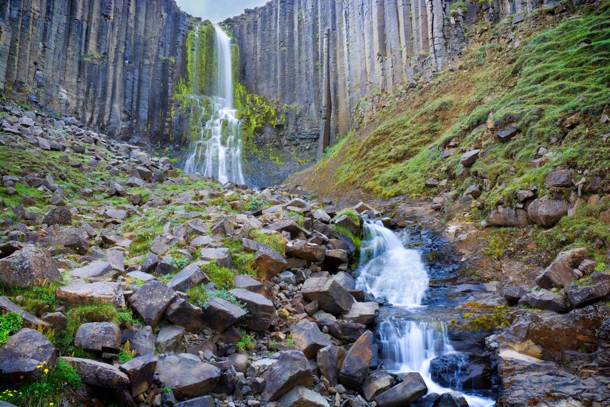 Waterfall cascading over dark basalt columns into a rocky, mossy landscape with a stream.