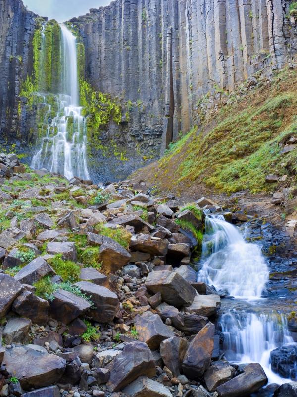 Waterfall cascading over dark basalt columns into a rocky, mossy landscape with a stream.