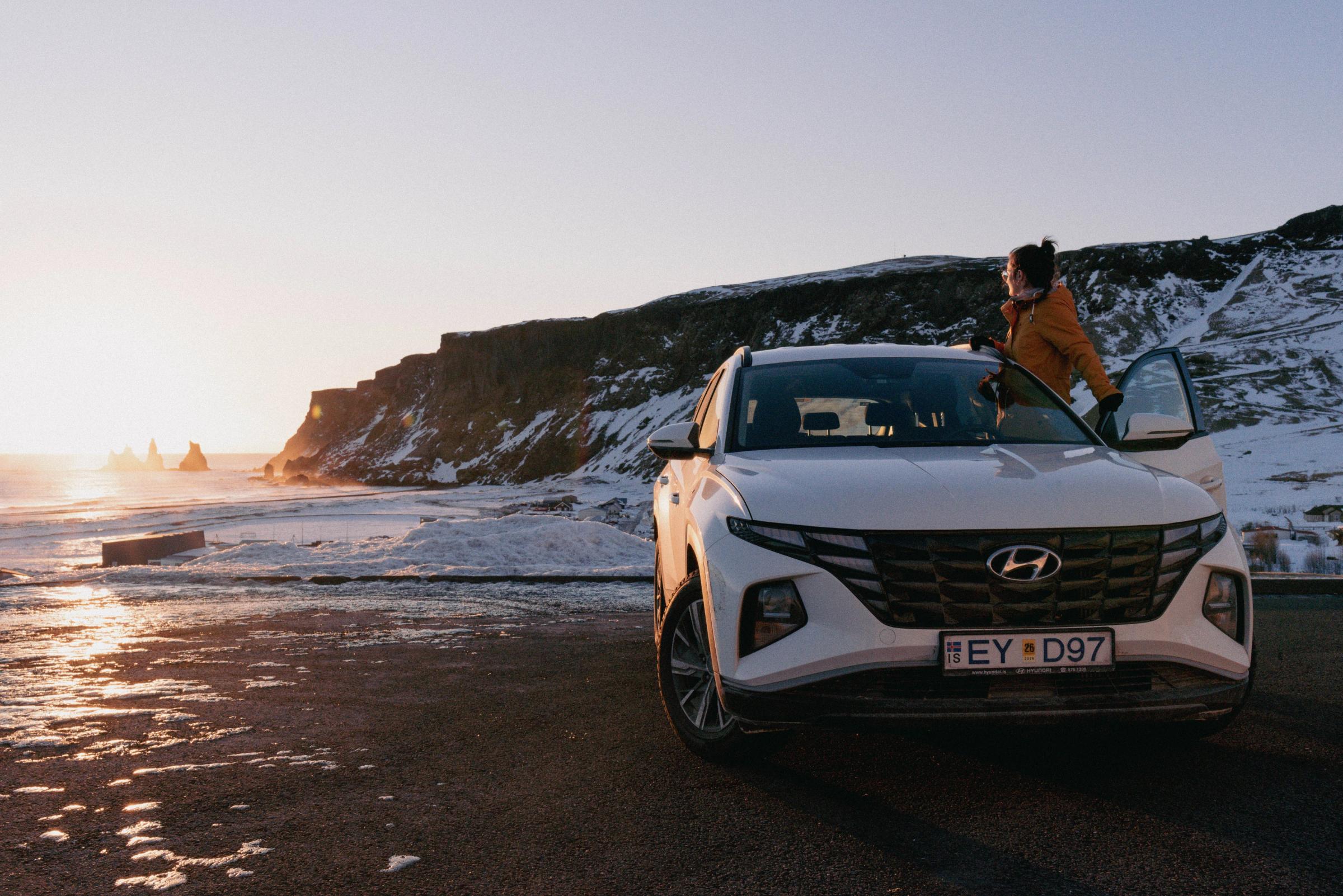 A white Hyundai SUV is parked on a snowy, icy coast at sunset, with a person leaning on it and sea stacks in the distance.