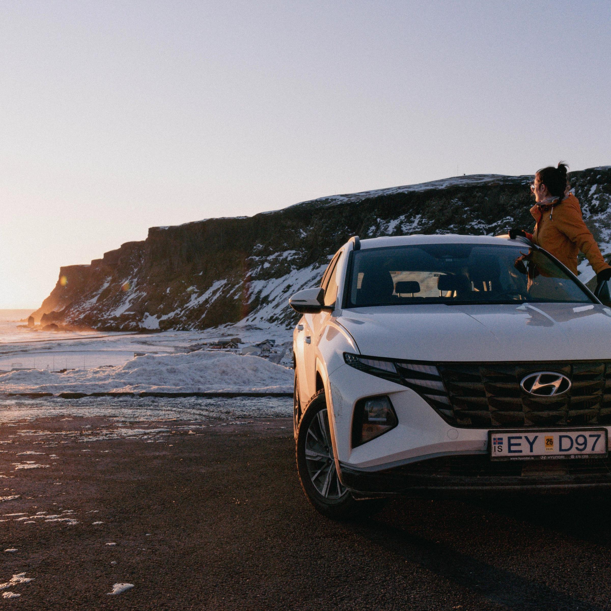 A white Hyundai SUV is parked on a snowy, icy coast at sunset, with a person leaning on it and sea stacks in the distance.