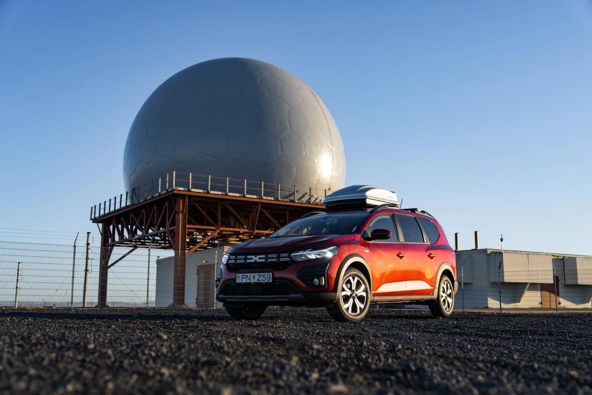 Coche de alquiler Dacia Jogger en Islandia, aparcado cerca de una estructura abovedada y equipado con un cofre de techo
