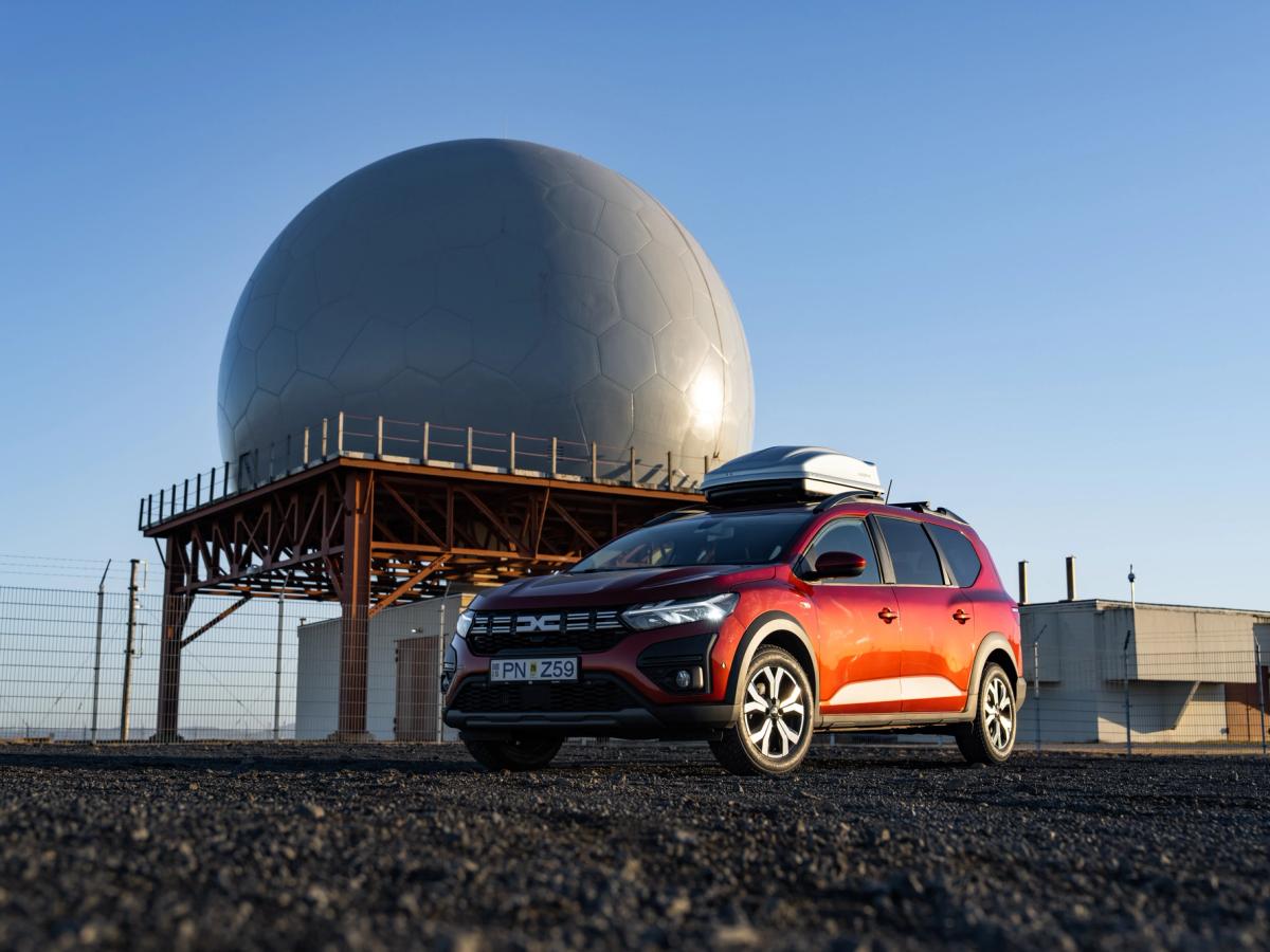 Fondo de monumento para Jogger Coche de alquiler Dacia Jogger en Islandia, aparcado cerca de una estructura abovedada y equipado con un cofre de techo