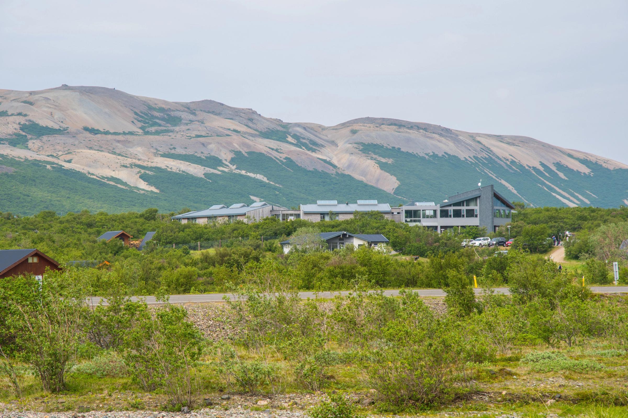 Edificios modernos y cabañas entre arbustos verdes y una carretera, con montañas al fondo.