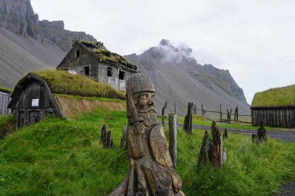 a wooden statue of a man in a helmet is sitting in the grass in front of a mountain at a viking village in iceland..