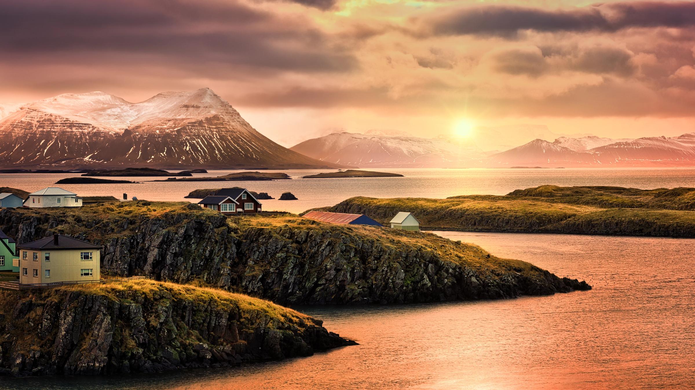 Coastal village with colorful houses on rocky islands, snow-capped mountains in the background, under a warm sunset sky.